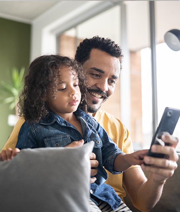 Image of man sitting on a couch with a child on his lap looking at a mobile phone screen